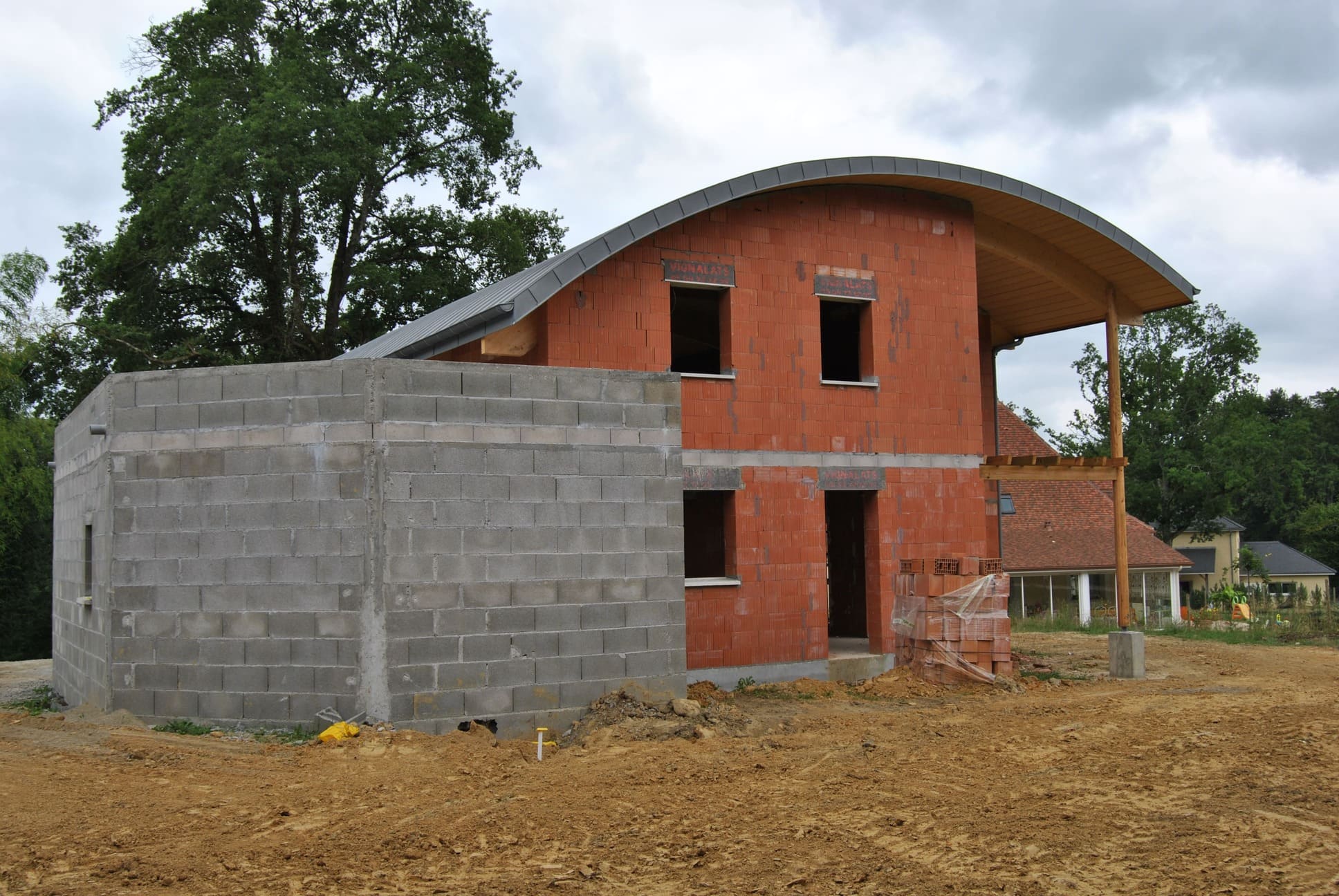 Photo d'un chantier de maçonnerie, on y voit une maison sur deux étages avec le toit arrondi et une charpente en bois. Une partie de la maison est en brique et sur un côté il y a une extension en parpaing gris.