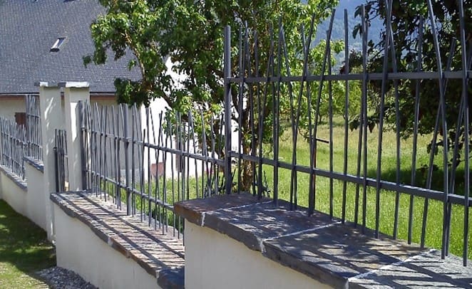 Photo d'une clôture de couleur gris foncé elles est posée sur une murette et est composée de barreaux entrecroisés. En arrière-plan on voit une pelouse et une maison au toit en ardoise.