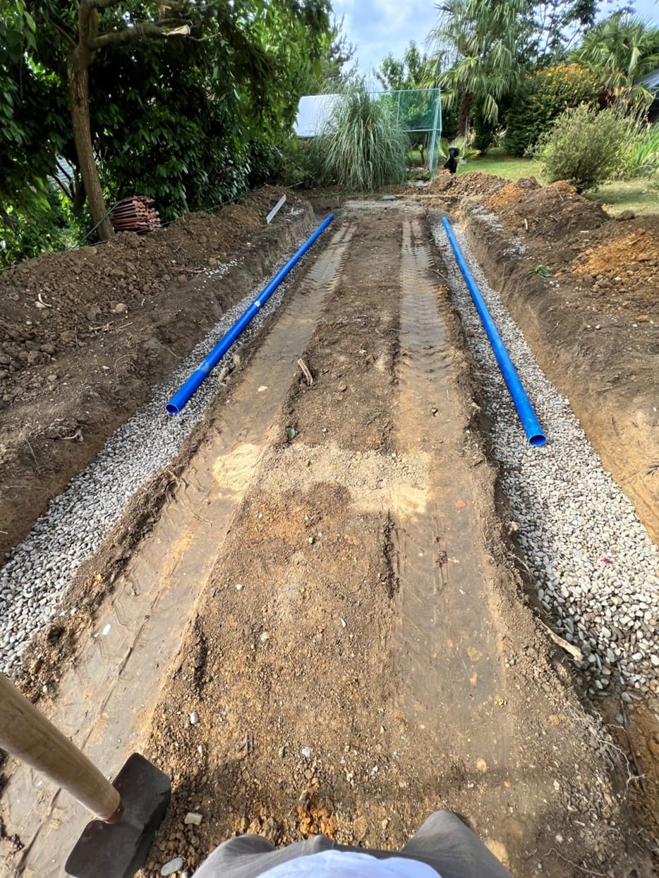 Photo d'un chantier, deux tranchées sont creusées dans un jardin et au fond de celles ci des graviers ont étés disposés et des gaines bleue sont posées dessus.