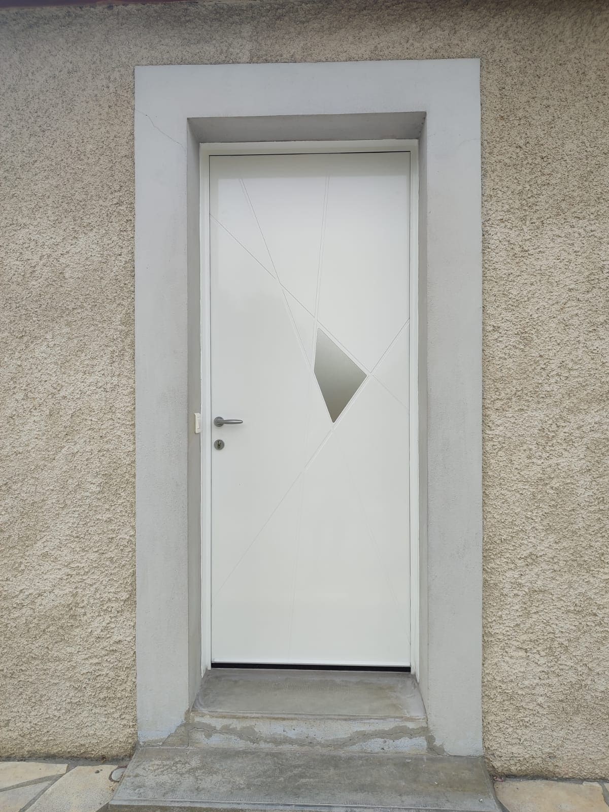 Photo d'une porte d'entrée blanche avec une poignée en aluminium, il y a un décor fait de quatre rainures qui se croisent formant un polygone gris en leurs centre.