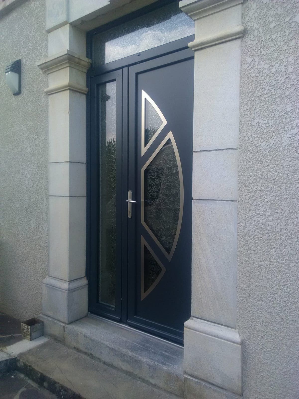 Photo d'une porte d'entrée gris anthracite avec une poignée en aluminium, il y a un décor fait de trois vitres en arrondi et entouré de métal. A gauche et au dessus de la porte des vitres laisse passer la lumière.